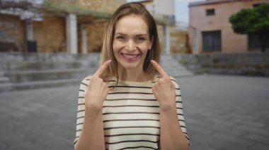 Yaşlı bir kasabanın kaldırım taşı sokağında iki işaret parmağıyla gülümseyen dudaklarını gösteren bir kadın; neşeli bir neşe.