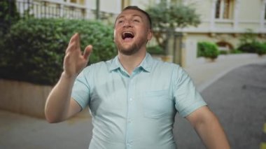 Adam ağızdan ağıza arıyor gözleri kapalı ve sakallı, açık mavi gömlek giyiyor, bir binanın yanındaki yapraklı bir sokakta duruyor; sevinç kutlaması.