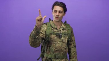 Kamuflaj üniformalı, künyeli ve taktik çantalı bir adam parmak tabancasıyla iki parmağını kaldırıyor. Stüdyonun mor arka planına yakın bir yerde..