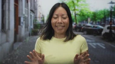 Sokakta, park halindeki arabaları ve yapraklı ağaçlarıyla kameraya doğru gülümseyen genç bir Latin kadın, sarı tişört giyiyor, arkadaş canlısı bir sevinç bağı var..