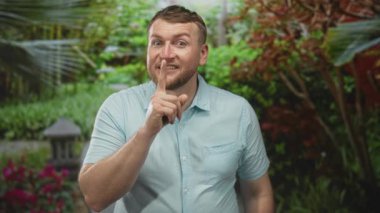 Yeşil yeşillik ve taş fenerle parktan dudaklara adam parmağı, açık mavi gömlek ve sakal gülümseme; şakacı gizli fener.