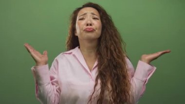 Avuç içleriyle omuz silken ve kaşlarını kaldıran, yeşil stüdyoda yüzü öne bakan ve pembe gömlekli bir kadın..