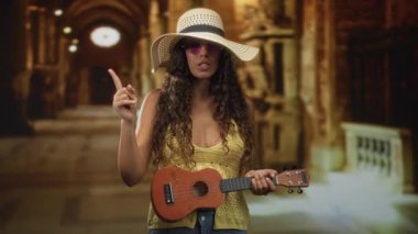 Kadın şarkı söylerken güneş şapkası ve yuvarlak güneş gözlüğü takan süslü bir bina koridorunda ukulele 'yi işaret ediyor ve tutuyor; neşeli seyahat merakı.