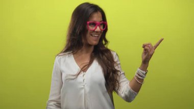 Kireç yeşili stüdyoda, pembe gözlük ve beyaz bluz takan kadın gülümserken parmağıyla sağa işaret ediyor..