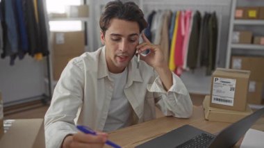 Adam telefonu kulağına tutuyor ve dizüstü bilgisayarın yanında kalemle yazıyor ve stüdyoda nakliye kutuları ve giyim raflarını istifliyor; girişimci odaklı.