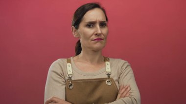 Kadın, kollarını kavuşturmuş kırmızı bir duvara karşı önlük takıyor ve izole edilmiş, canlı bir ortamda güven ve bağımsızlığını ifade ediyor..