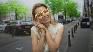 Sarı gözlüklü kadın gözlerini kapatıyor ve park halindeki arabalarla dolu hareketli şehir caddesinde elleri yanağına kenetlenmiş halde duruyor..