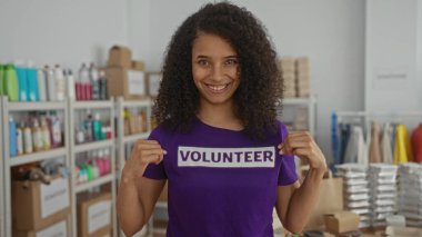 Bağış merkezinde gönüllü gömlekle gülümseyen bir kadın, arka planda çeşitli eşyalar, kapalı alanlarda aktif katılımı gösteriyor..