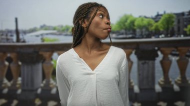 Süslü saçlı bir kadın beyaz bir bluz içinde dudaklarını büzüştürüyor Amsterdam 'da kanal üzerinde bir sokak köprüsünde; oyuncu bir merak.