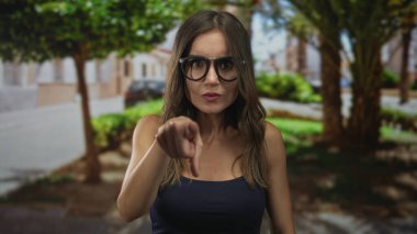 Latin kökenli genç bir kadın, yeşil ağaçlar ve park edilmiş arabalarla çevrili caddedeki izleyiciyi işaret ediyor..