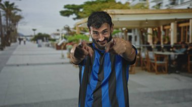 Mavi futbol üniformalı adam kamerayı caddede gösteriyor, sakalı görünüyor ve ön planda işaret ediyor; oyuncu..