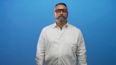 Mavi stüdyoda gözlük takan gri sakallı ve kapalı gözlü orta yaşlı İspanyol bir adam..