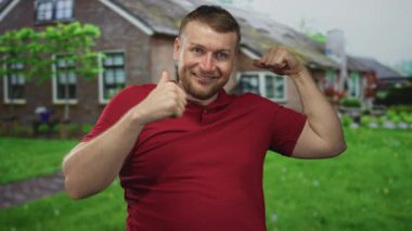 Adam bina girişinde kollarını esnetiyor kırmızı gömlekli kameraya doğru gülümsüyor kaslarını ve kaslarını gösteriyor; güven kutlaması.