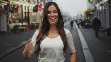 Kadın kalabalık bir şehir caddesinde kamerayı işaret ediyor, kutlama için yumruğunu kaldırırken geniş bir gülümseme; kendine güven.