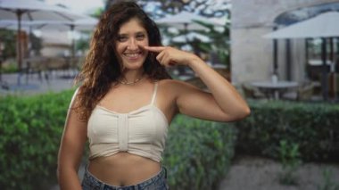 Kadın parmaklarını yanağına doğrultur ve taştan bir binanın yanındaki midriff 'i kameraya bakıp gülümserken gösterir..