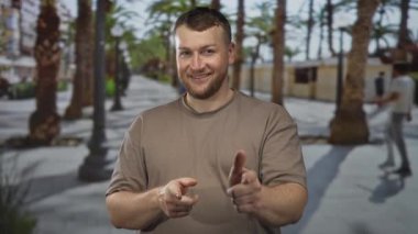 Adam gülümsüyor ve her iki işaret parmağı kamera üzerinde güneşli bir palmiye ağacı çizgi gezinme caddesinde bulanık geçen ile; oyuncu selamlama.