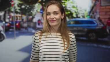 Çizgili tişört giyen kadın, şehrin güneşli caddesinde hafifçe eğilmiş başıyla gülümsüyor..