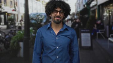 Kıvırcık saçlı bir adam işlek bir şehir caddesinde kolları önkollu gözler, mavi gömlek ve yuvarlak gözlük giyiyor ve nazikçe gülümsüyor; sakin tefekkür.