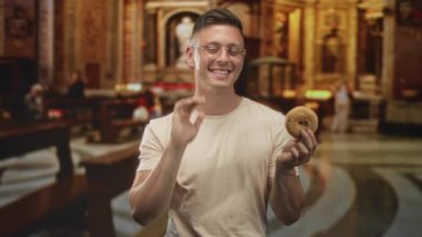 Gözlüklü adam donutu sağ elinde tutuyor, gülümsüyor ve başını eğiyor süslü binanın içinde; eğlenceli bir merak.