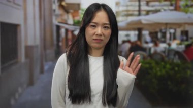 Bir kadın kalabalık bir sokak terasında insanların açık şehir ortamında bir restoranda yemek yemesiyle el sallıyor..