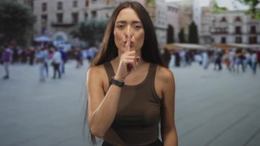 İşlek bir şehir meydanında sessiz kalmak için parmakları dudaklarına kadar uzanan genç İspanyol kadın, kahverengi kolsuz tişört ve şık kıyafetler giyiyor, uzun saçlar görünür; sessizlik gizlilik.