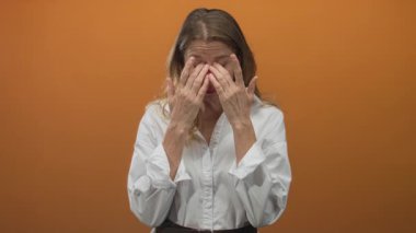 Stüdyoda ellerini gözlerine bastıran bir kadın, turuncu duvarla burun köprüsünü ovuyor, beyaz gömlek giyiyor, yorgunluk tedavisi görüyor..