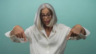 Orta yaşlı gri saçlı kadın işaret parmağıyla işaret ederek ellerini gösteriyor ve camgöbeği atölyesinde beyaz gömlek giyiyor..