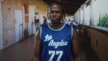 Adam kol aşağı bakarken mavi Los Angeles forması ve bileklik giyerek bina içinde kaslarını esnetiyor..