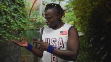 Genç adam, yeşil yeşillikler arasında parkı işaret ederek ABD spor forması ve mavi bileklik takıyor..