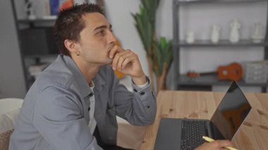 Genç adam rahat bir oturma odasında dizüstü bilgisayar kullanırken gitar ve bitkilerin yanında düşünceli bir yüz ifadesi gösteriyor..