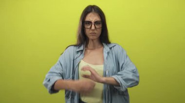 Kireç yeşili bir stüdyoda kolları çapraz bir kadın düz bir duvara karşı, gözlük ve kot gömlek giyiyor; sinir bozucu sınır.
