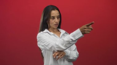 Kırmızı stüdyoda eli çapraz beyaz gömlekli bir kadın kamerayı kapalı olarak işaret ediyor; iddialı..