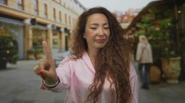 Kadın, alışveriş merkezleri, ekiciler ve yayaların bulunduğu işlek bir caddede kamerayı işaret ediyor; özgüven odaklı.