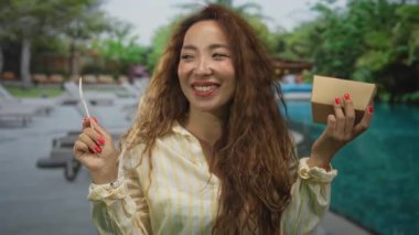 Kadın havuz başında karton kutu ve ahşap çatal tutuyor, gülümsüyor ve kırmızı tırnaklarını gösteriyor; mutlu yaz tatili.