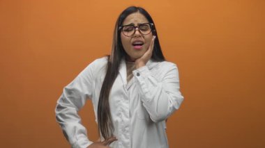 Kadın doktor, beyaz önlüklü ve gözlüklü genç bir kadın kremle el ele tutuşuyor, turuncu stüdyoda diş ağrısı çekiyor..