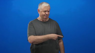 Stüdyoda, koyu renk t-shirt giyen, mavi arka planda sessiz kalan, parmaktan dudağa şşş yapan son sınıf öğrencisi..