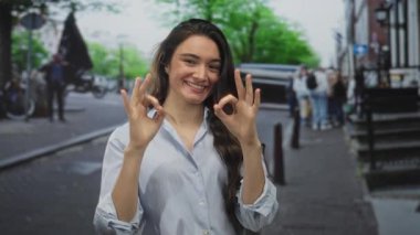 Genç İspanyol kadın gülümsüyor ve el işareti yapıyor. Meşgul şehir caddesinde dükkan vitrinleriyle sıralanmış dururken. Onay güven pozitifliği güçlendirici..