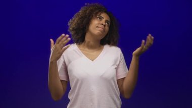 Kadın iki elini de kaldırır, avuçlarını kaldırır ve açık mavi bir stüdyoda soluk pembe bir tişört giyer, yukarı bakar, hayal kırıklığı.