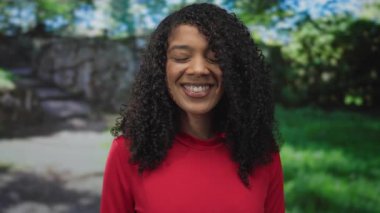 Ormanda kırmızı gömlekli bir kadın parlak güneş ışığı altında taş duvarın yanında gülümsüyor. Enerji mutluluğu ısıtıyor..