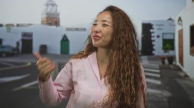 Kadın, beyaz bina ve yaya geçidi önündeki sokakta, görünür elleri ve sıcacık bir gülümseme ile başparmağıyla işaret ediyor; neşeli iletişim.