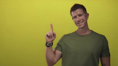 Sarı stüdyoda çıplak elle işaret eden adam zeytin tişört ve bilezik takıyor; dost canlısı..