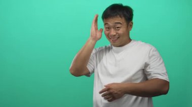 Genç Çinli adam yeşil stüdyoda elleri ile çerçeve gülümsüyor, doğrudan objektife bakıyor; merak hayal gücü yaratıcılığını hayrete düşürüyor.