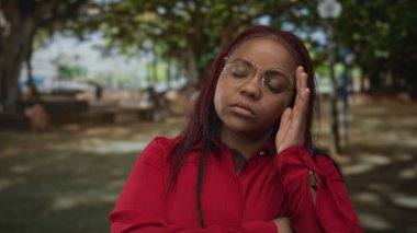 Kırmızı bluz giyen ve gözlük takan kadın çenesini eliyle tutuyor ve yapraklı ağaçların altında kollarını kavuşturuyor..