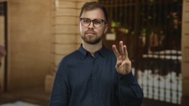 Gözlüklü adam üç parmağını kaldırır, öne doğru tutar ve gün ışığında bina girişinde avuç içi teklif eder; düşünceli davet..