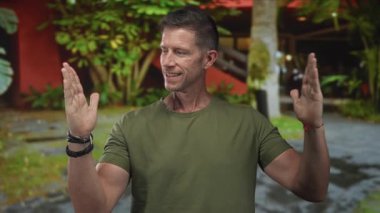 Avuç içi açık bir adam bir binanın ve yapraklı avlunun önünde jest yapar; sakin farkındalık dengesi.