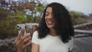 Kadın akıllı telefonu kol mesafesinde tutuyor ve yeşil şehir parkında güneş ışığı altında iki parmakla barış jesti yapıyor..