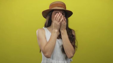 Hasır şapka giyen genç İspanyol kadın stüdyoda sarı arka plana karşı iki eliyle burnunu çimdikliyor..