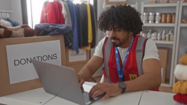Bağış kutusunun yanında dizüstü bilgisayarda yazan kıvırcık saçlı bir adam gönüllü halat ve kırmızı yelek giyiyor..