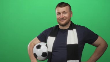 Yeşil stüdyoda elleri kalçasında futbol topu tutan, gülümseyen, siyah-beyaz atkı takan ve donanma tişörtü giyen bir adam..