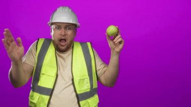 Beyaz kasketli ve yüksek görüş mesafesi olan yelekli bir adam, mor stüdyoda ağzının üzerinde yeşil elma tutuyor; sürpriz güvenlik endişesi.